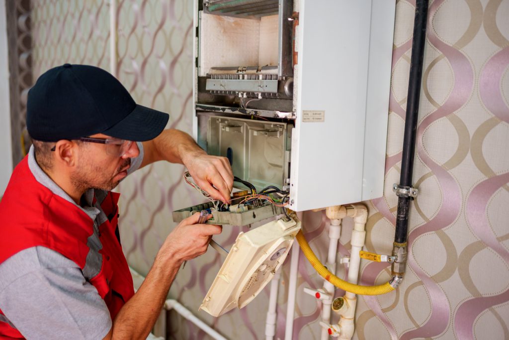 A technician in a red vest and hat carefully works on the internal components of a wall-mounted boiler