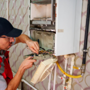 A technician in a red vest and hat carefully works on the internal components of a wall-mounted boiler