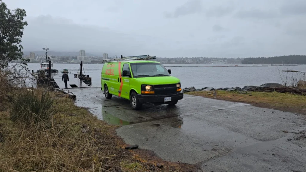 Bright green service van parked beside a small ferry ramp on a rainy waterfront, with a city skyline across the water