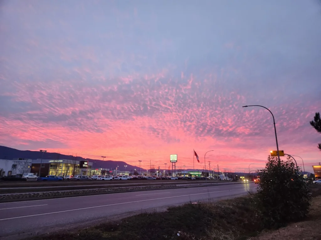 Pink sunset over a commercial roadway with mountains, storefronts, cars, and streetlights